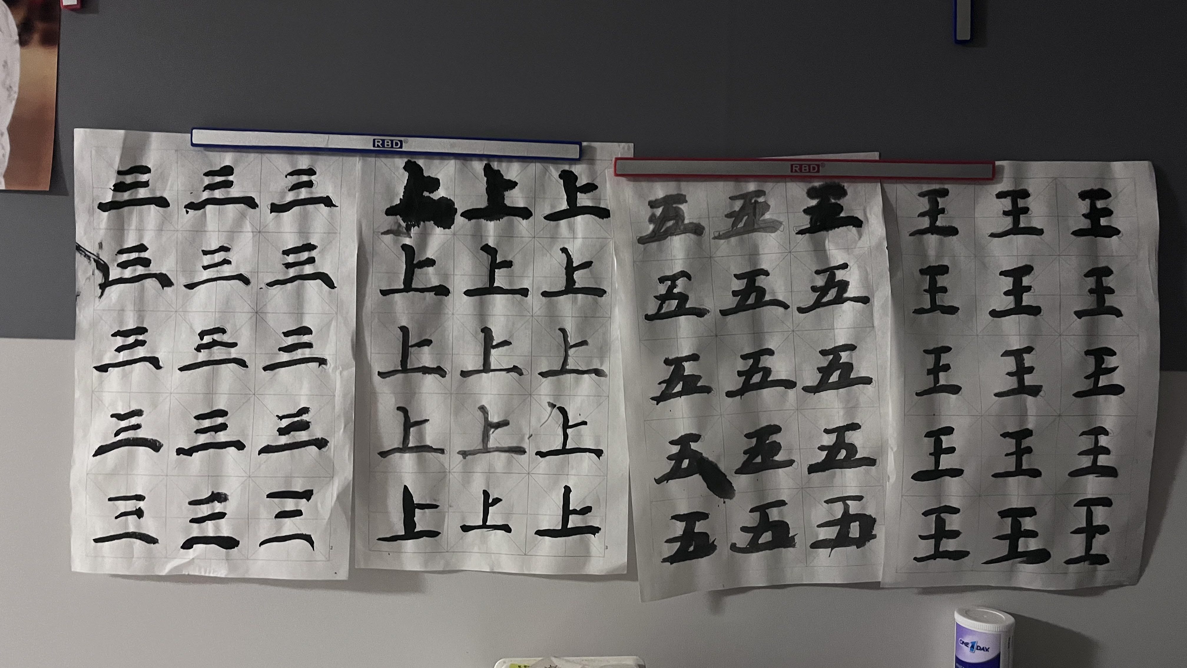 Three sheets of paper displaying Chinese characters. The left sheet primarily features the character for 'three' (三), while the center sheet shows the character for 'up' (上). The right sheet displays the character for 'five' (五).